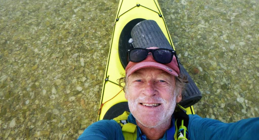 A person leans back while sitting in a yellow kayak to take a selfie. Below the boat, you can see clear water, showing the rocks below. 